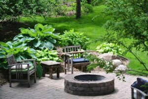 An outdoor patio with a brick fire pit, wooden chairs, a table, and green foliage.